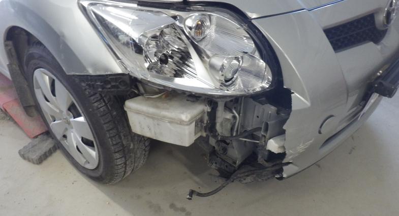 Hopeanharmaan auton etukulman vaurio kolarin jäljiltä, jossa näkyy rikkoutunut puskuri ja paljastuneita osia.