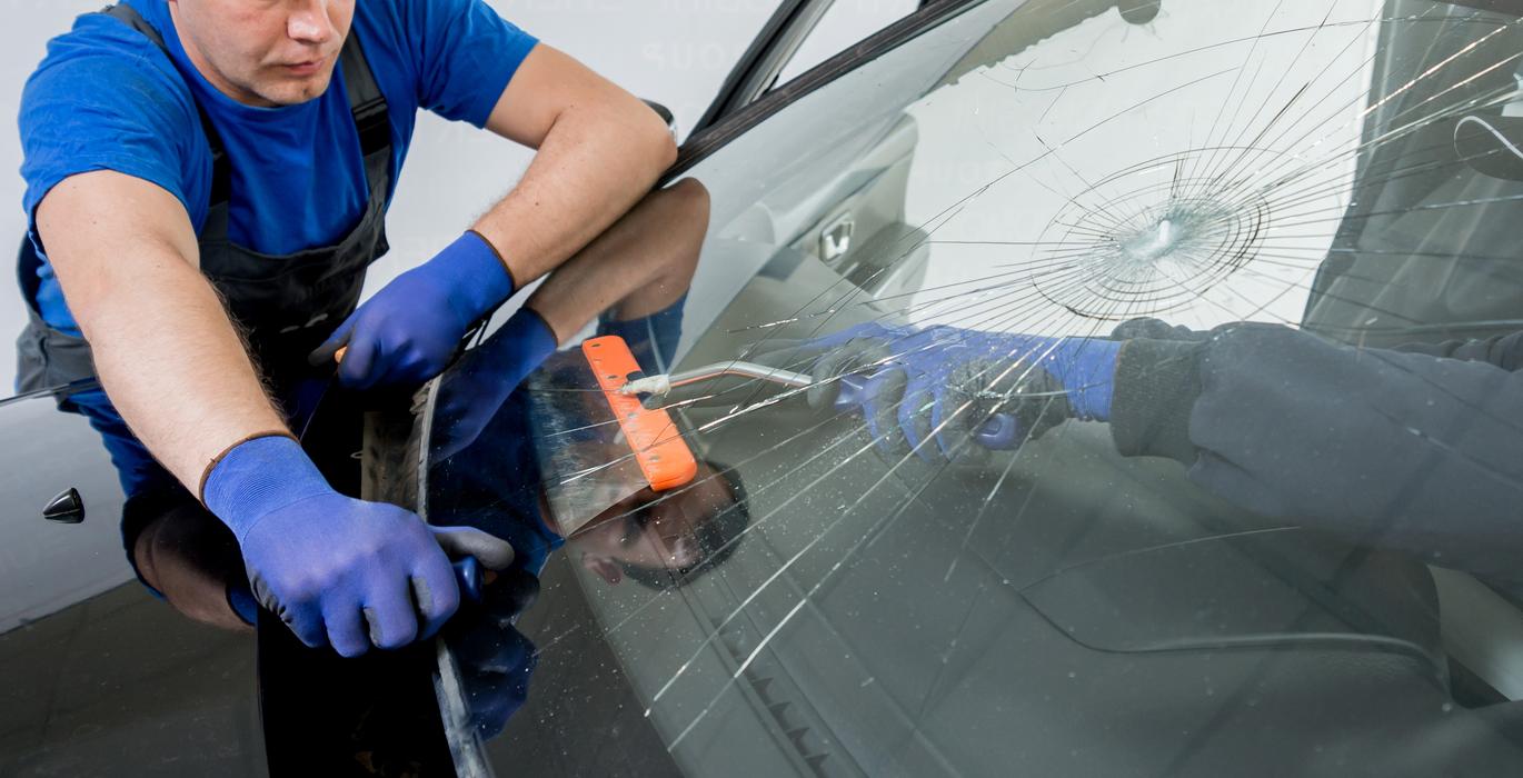 Automekaanikko vaihtamassa säröytynyttä tuulilasia siniset suojakäsineet kädessä.